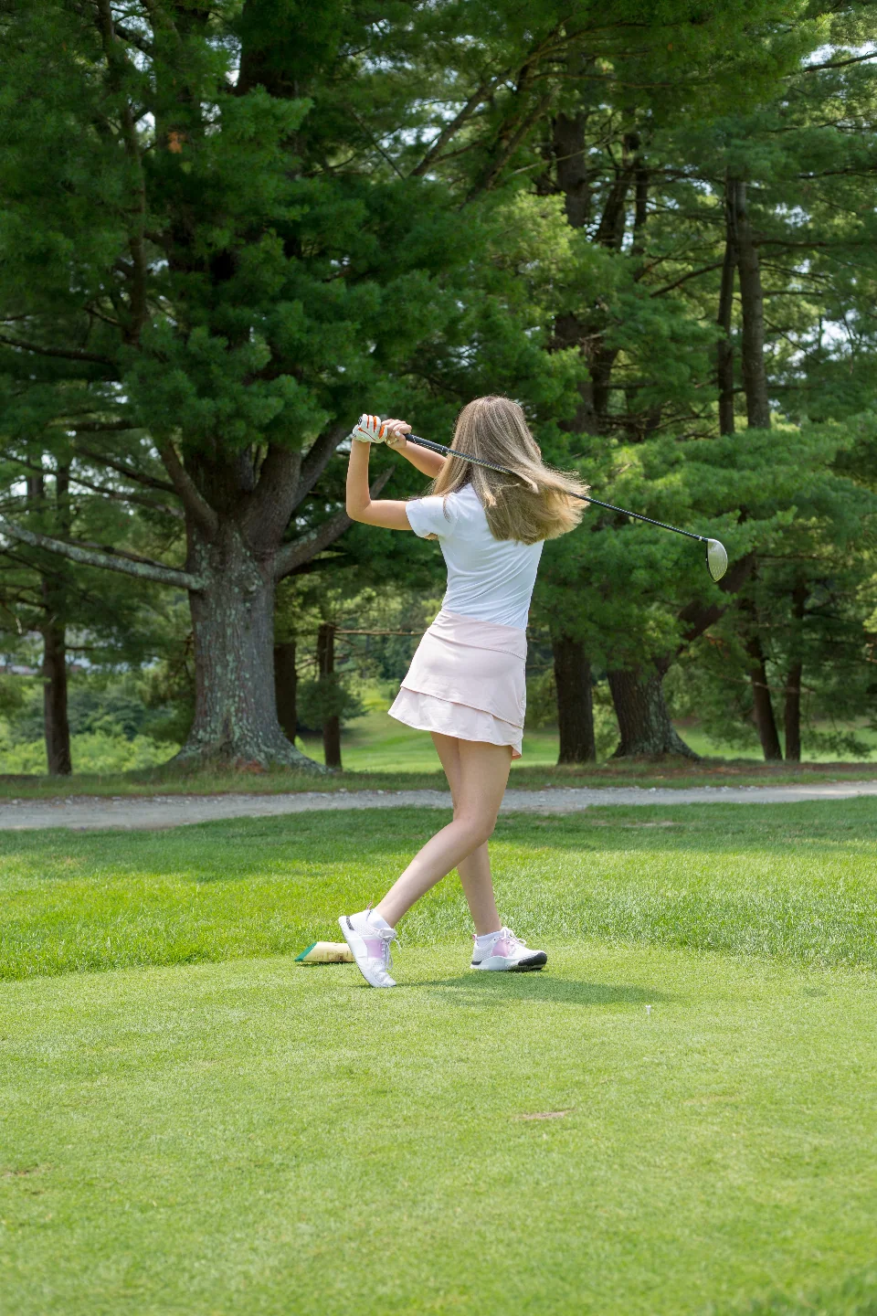 A golfer in a white polo follows through on her tee shot, tall pines behind her
